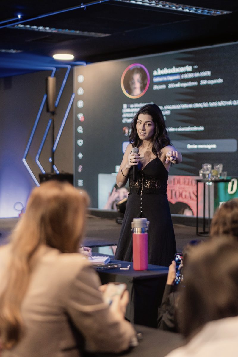 Kath Zaparoli no palco apontando para a plateia, com o perfil da Bobo da Corte no telão atrás dela