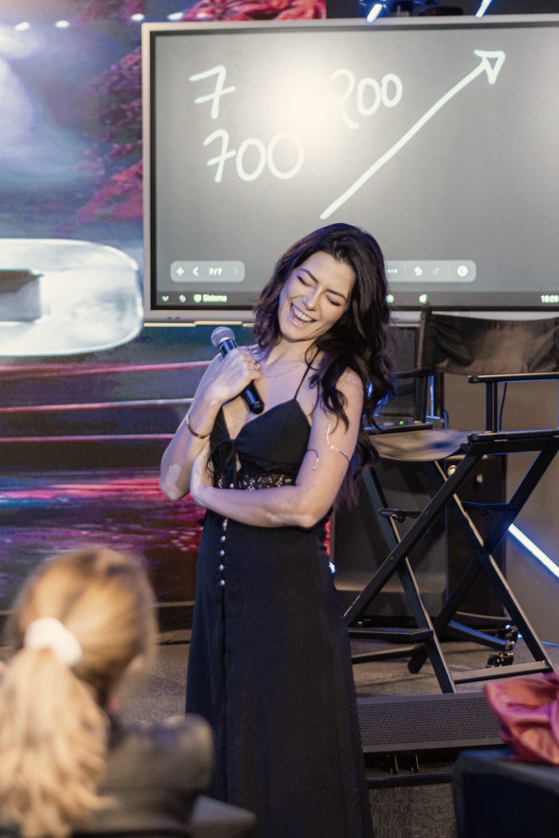 Kath Zaparoli rindo no palco, com o microfone junto ao peito